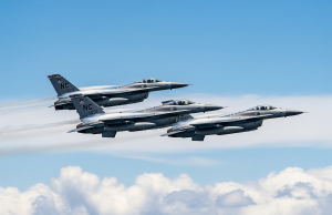 Trīs F-16 tipa iznīcinātāji pelēkā krāsā lido formācijā virs mākoņiem skaidrās, gaiši zilas debesīs