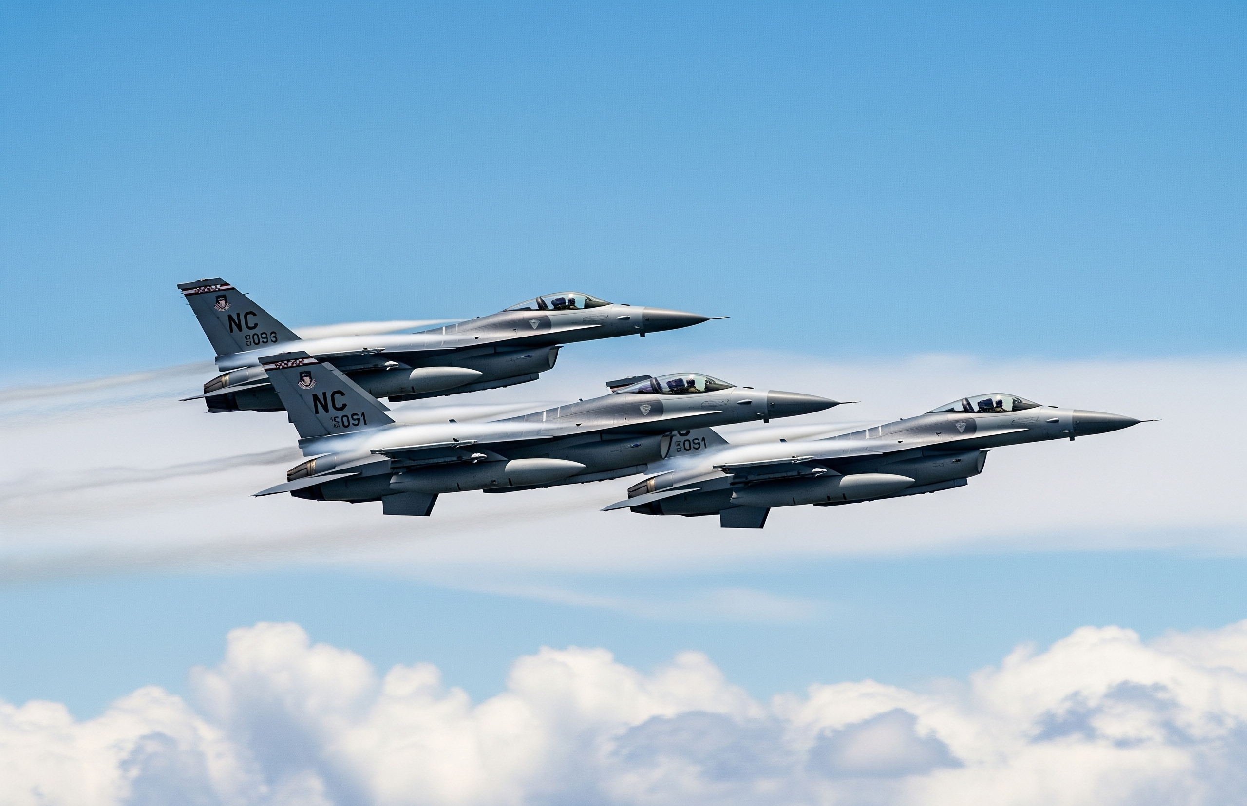 Trīs F-16 tipa iznīcinātāji pelēkā krāsā lido formācijā virs mākoņiem skaidrās, gaiši zilas debesīs