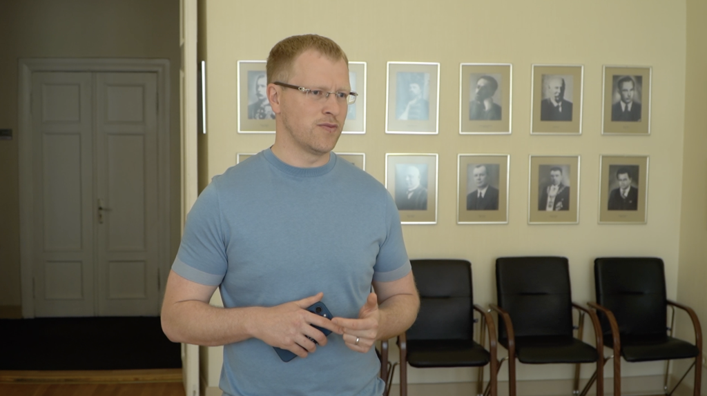 Vīrietis ar brillēm gaišā t-kreklā (Andrejs Elksniņš) runā telpā ar portretiem pie sienas