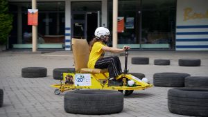 Meitene ar ķiveri brauc ar pašbūvētu dzeltenu transportlīdzekli starp riepu šķēršļiem tehnoloģiju festivālā