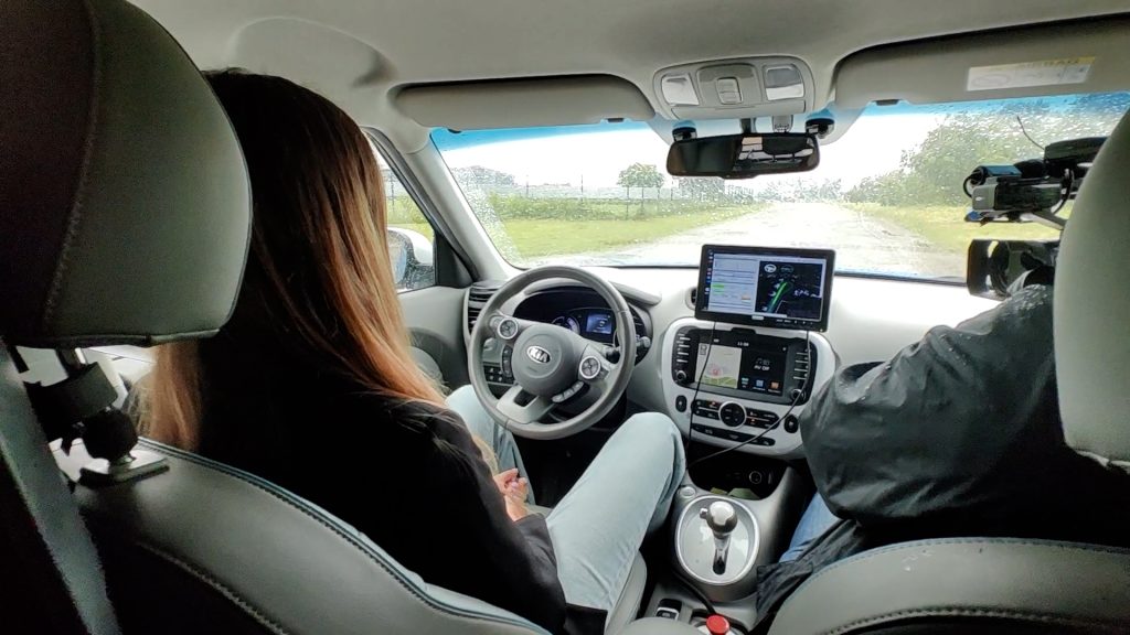 Sieviete sēž pasažiera sēdeklī pašbraucošā auto ar planšetdatoru uz paneļa, kas rāda navigācijas informāciju.