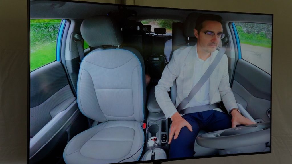 Ekrāns, kurā redzams vīrietis sēžot pasažiera sēdeklī pašbraucošā auto, ar drošības jostu un rokām atstutētām uz ceļgaliem