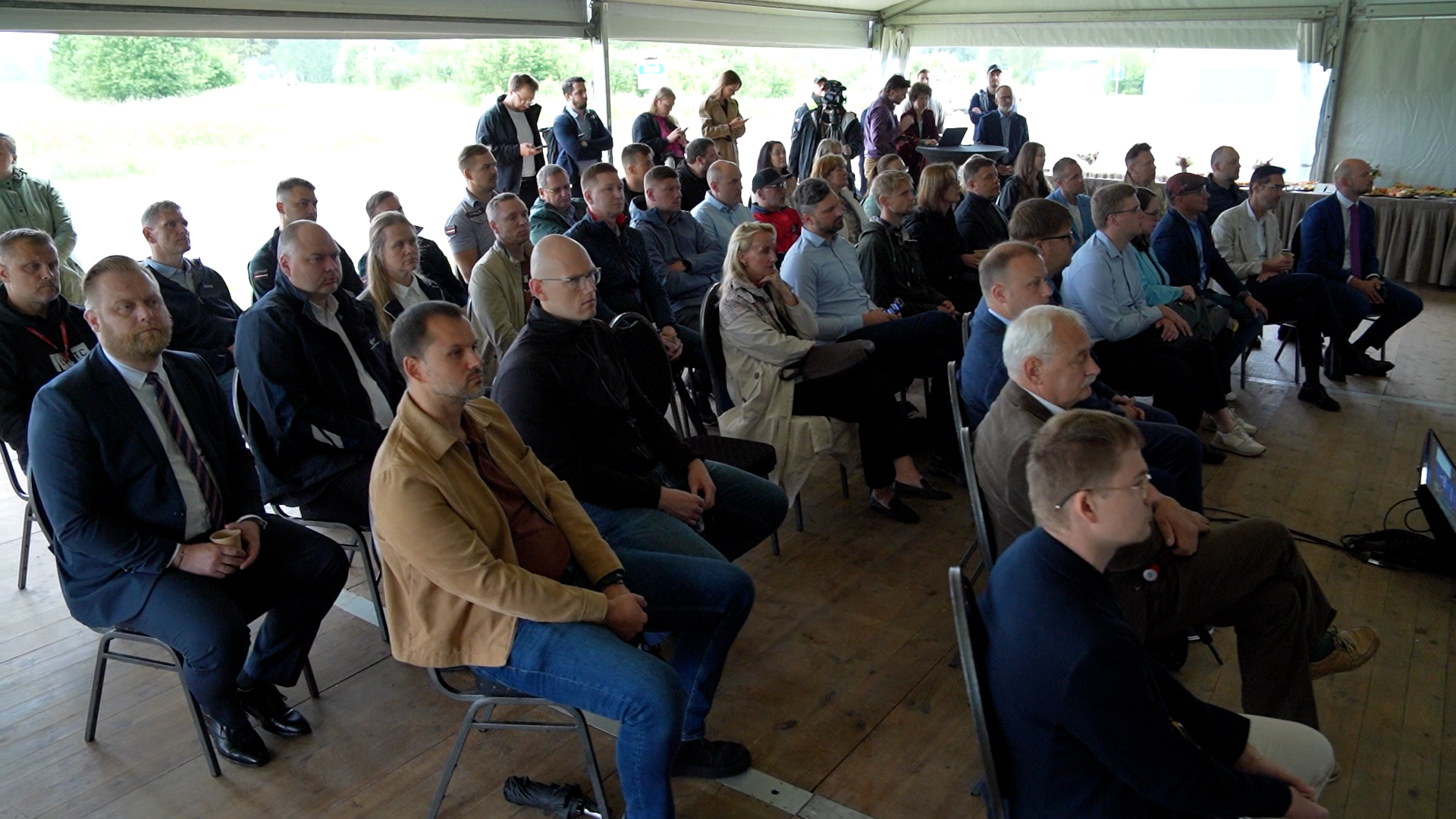 Liela auditorija, kas sastāv no vīriešiem un sievietēm, sēž krēslos un klausās prezentāciju