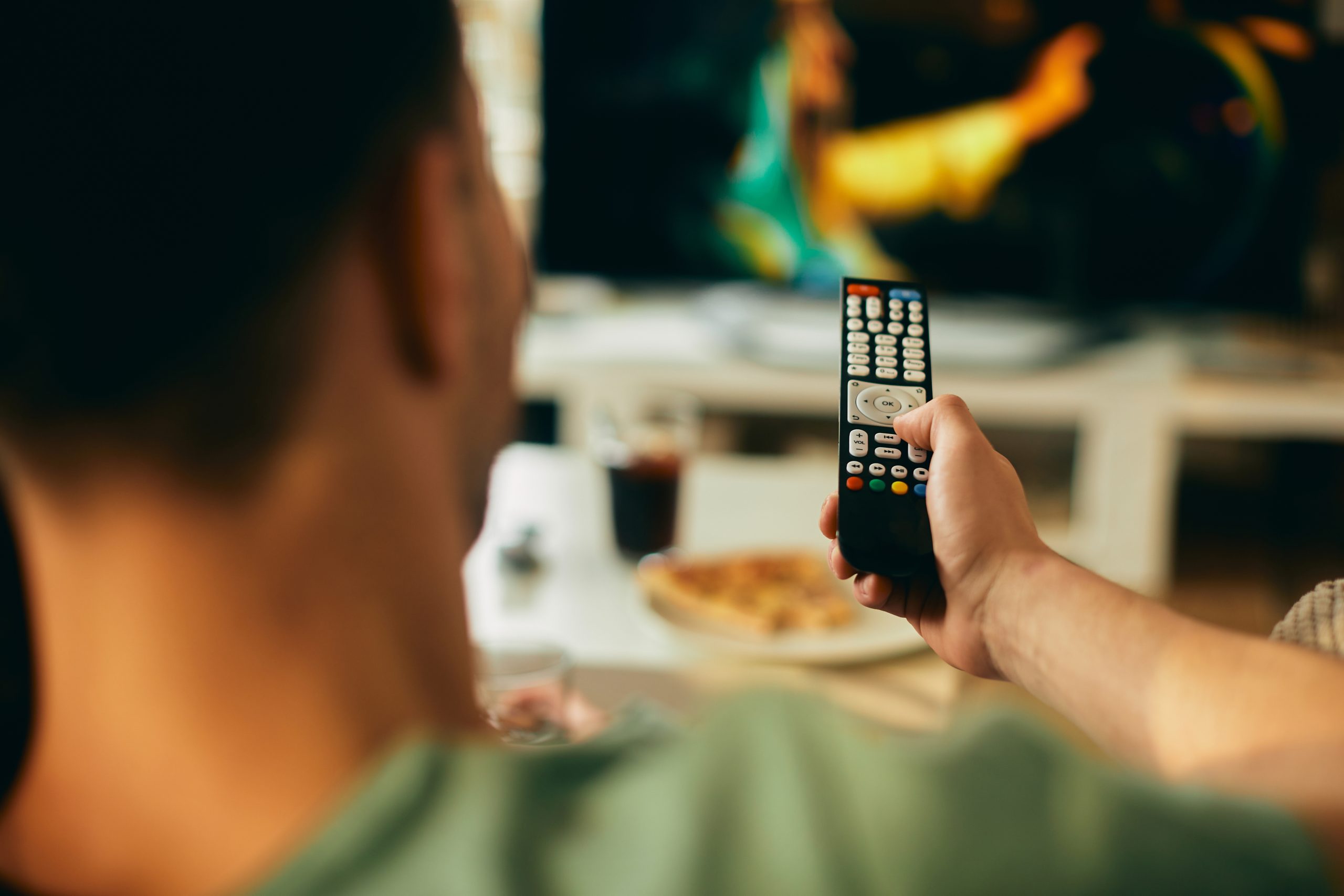 Cilvēks mājās skatās televizoru, turot rokās pulti.