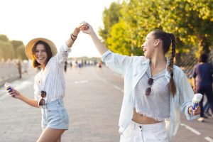 Divas jaunas sievietes priecīgi dejo ārā saulainā dienā, turot saldējumu un smejoties