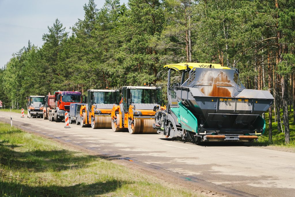 Rindā izvietotas ceļu būves mašīnas, tostarp asfalta ieklājējs un veltņi, gatavībā darbam pie meža