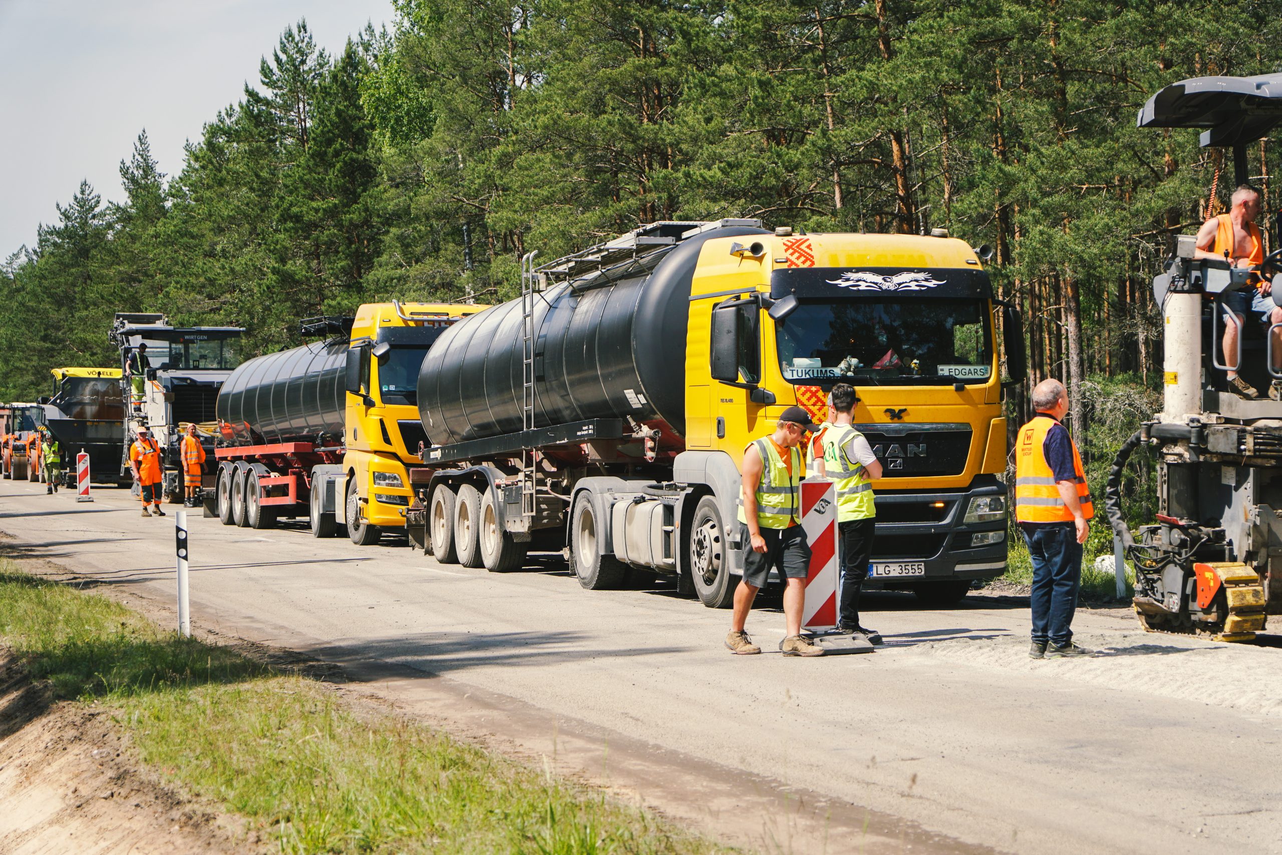 Trīs dzeltenas kravas automašīnas ar cisternām rindā uz ceļa, blakus stāv strādnieki.