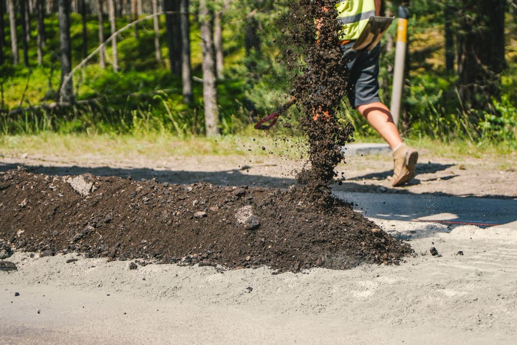 Strādnieks melno graudaino materiālu ber uz ceļa, veidojot kaudzi, ar mežu fonā