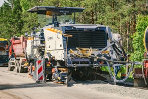 Liela ceļu būves mašīna "WIRTGEN" darbojas uz ceļa, blakus tai stāv kravas automašīna