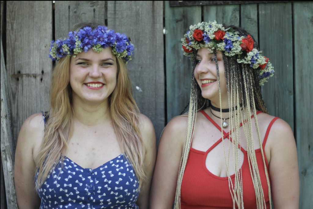 Divas smaidošas sievietes ar pavasara ziedu vainagiem galvās, stāvot pie vecas koka sienas