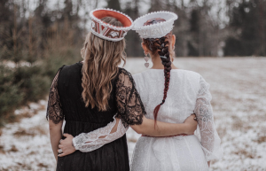 Divas sievietes tradicionālos tērpos vai stilizētos apģērbos, ar galvā kroņiem, stāv sadevušās rokās uz sniegota fona