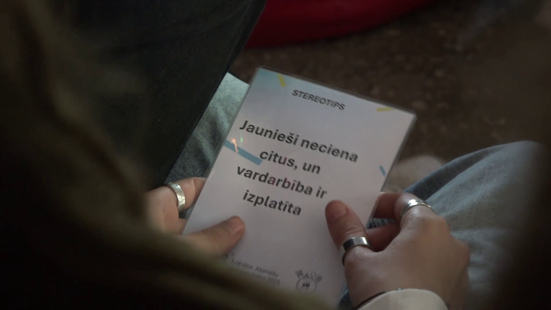 uvplānā redzams plakāts ar uzrakstu "Jaunieši neciena citus, un vardarbība ir izplatīta".