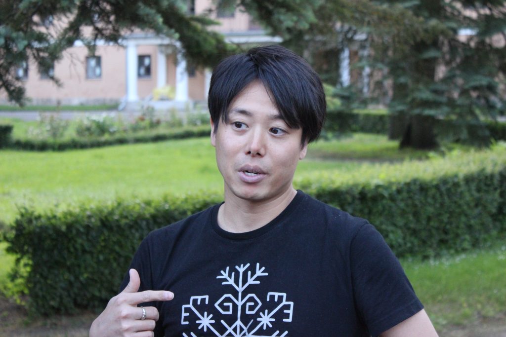 Masaki Nakagava, jauns vīrietis ar tumšiem matiem, tērpies melnā T-kreklā ar gaišu ornamentu, runā āra apstākļos ar zaļu zāli un krūmiem fonā