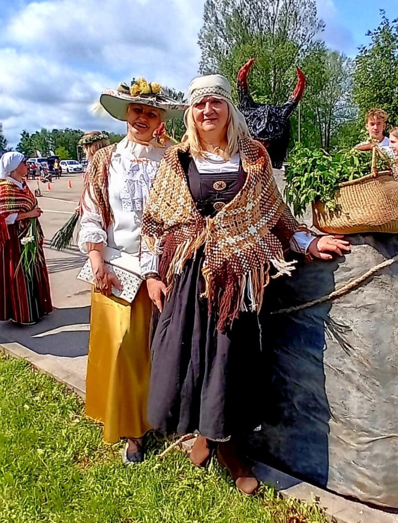 Divas sievietes tautas tērpos vai stilizētos apģērbos, viena ar platu cepuri, otra ar adītu lakatu, stāv ārā, fonā redzams "velna" tēls