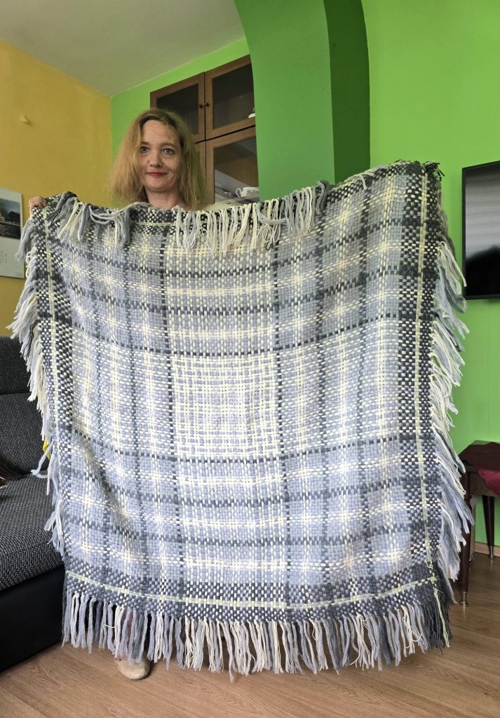 Sieviete ar gaišiem matiem, smaidot tur rokās lielu, pelēki-baltu rūtainu lakatu ar bārkstīm, stāvot telpā