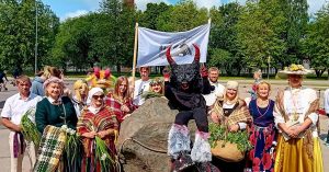 Grupa cilvēku, tostarp bērni, dažādos krāsainos tērpos un mītisku tēlu kostīmos, stāv pie liela akmens ar baltu karogu fonā, priecīgi pozējot kamerai