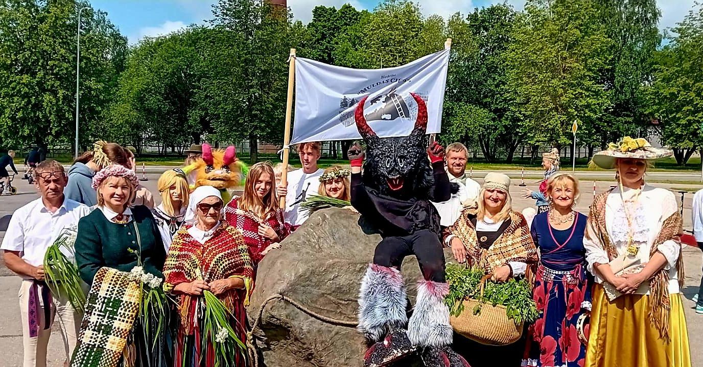 Grupa cilvēku, tostarp bērni, dažādos krāsainos tērpos un mītisku tēlu kostīmos, stāv pie liela akmens ar baltu karogu fonā, priecīgi pozējot kamerai