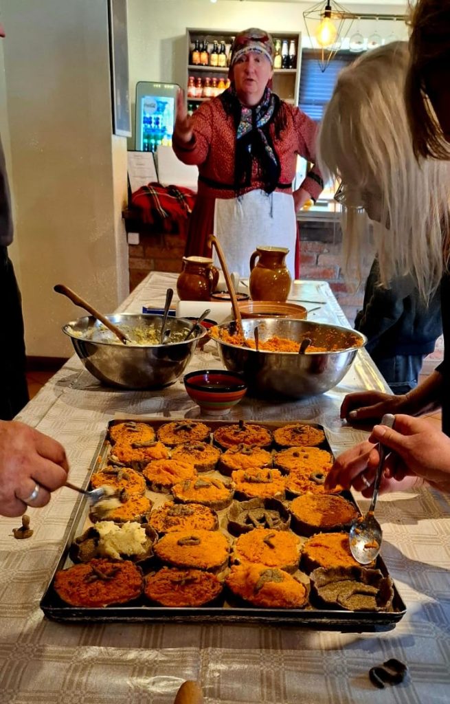 Sieviete tradicionālā tērpā stāv pie galda ar daudziem sklandraušiem un citām ēdienu bļodām, demonstrējot kulinārijas meistarklasi