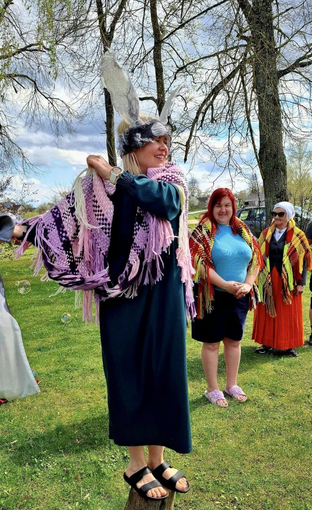Sieviete ar liela zaķa ausīm uz galvas un violetu šalli plecos stāv uz koka bluķa, aiz viņas divas citas sievietes, visas brīvā dabā