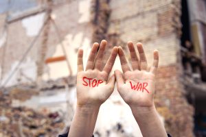 Roku plaukstas ar uzrakstu "STOP WAR" sagrautas ēkas fonā, simbolizējot pretestību karam.