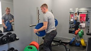 Vīrietis sporta zālē uzliek svara ripas uz stieņa