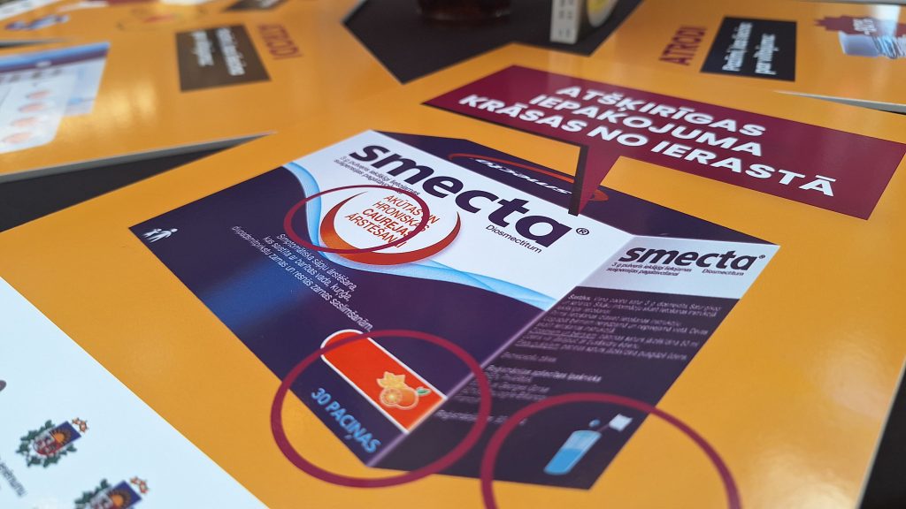 Tuvplānā redzams informatīvais materiāls par viltotu "Smecta" iepakojumu, ar iezīmētām atšķirībām.