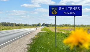 Ceļa zīme ar uzrakstu "Smiltenes novads" un tā ģerboni, kas norāda uz novada robežu pie autoceļa, ko ieskauj lauki un meži
