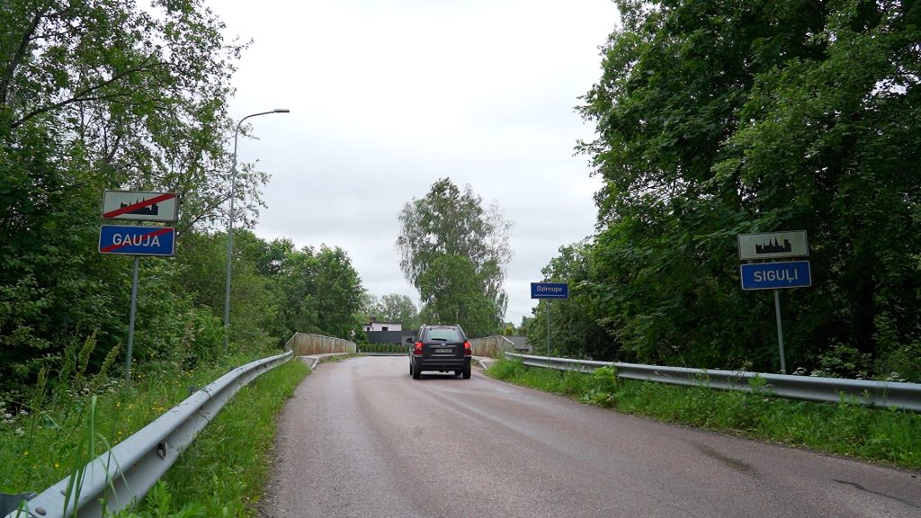 Automašīna brauc pa ceļu, kas ved uz Gaujas tiltu, ar zīmēm "Gauja" un "Siguli"