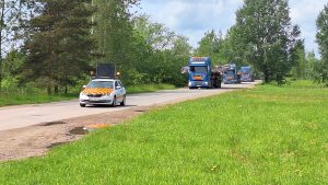 Speciālais pavadošais transports ar dzeltenām un oranžām svītrām priekšplānā, un aiz tā garš smagsvara sastāvs ar vēja turbīnu torņa konstrukcijām uz ceļa ar kokiem abpus