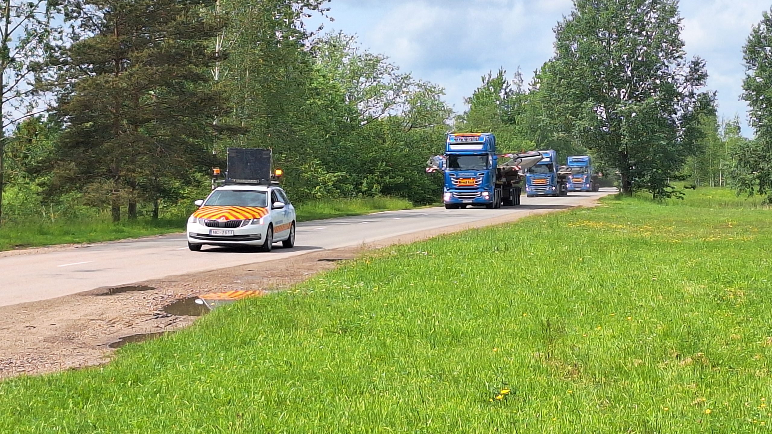 Speciālais pavadošais transports ar dzeltenām un oranžām svītrām priekšplānā, un aiz tā garš smagsvara sastāvs ar vēja turbīnu torņa konstrukcijām uz ceļa ar kokiem abpus