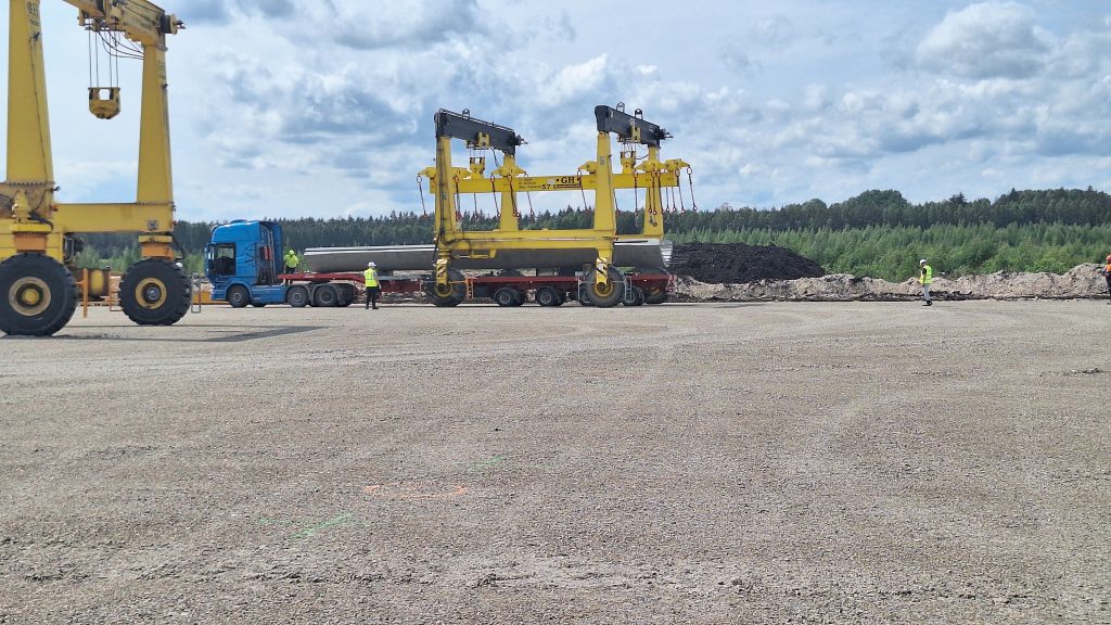 Liels, dzeltenas krāsas celšanas portālceltnis, kas gatavojas izkraut vēja turbīnas konstrukciju uz grants laukuma