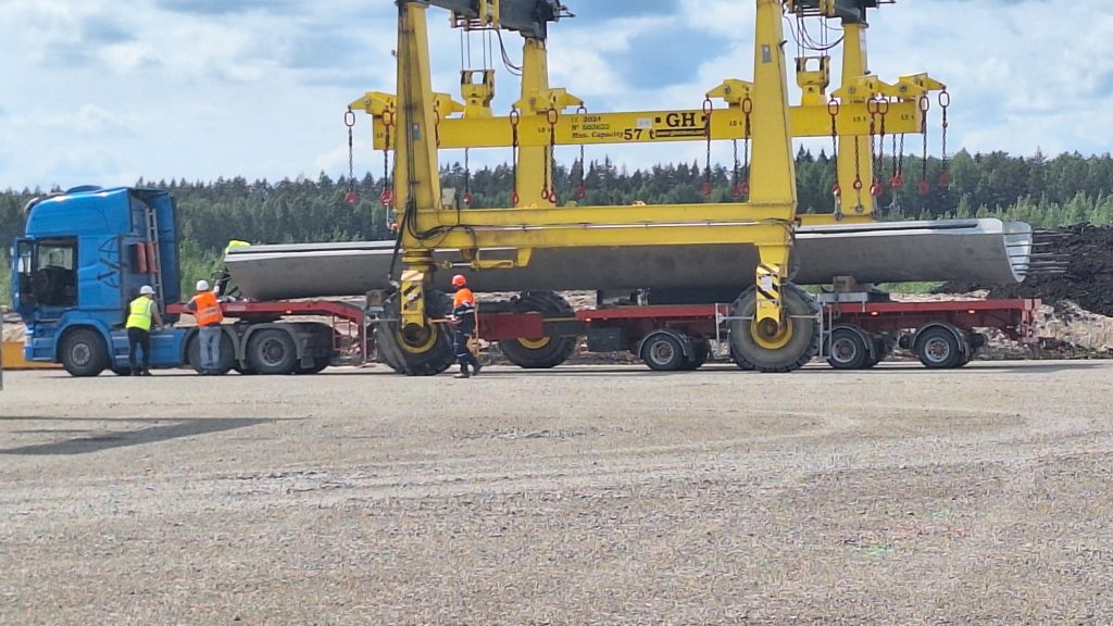 Liels dzeltenas krāsas celšanas portālceltnis, kas izkrauj vēja turbīnas konstrukcijas sekciju no transporta piekabes