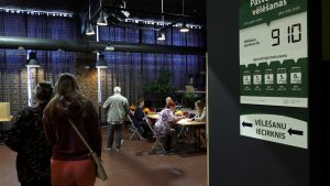 Vēlēšanu iecirknis ar cilvēkiem un informatīvu afišu par 2025. gada vēlēšanām