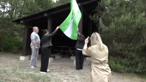 Cilvēki pacel zaļo “NaaC” pieejamības karogu pie meža nojumes.