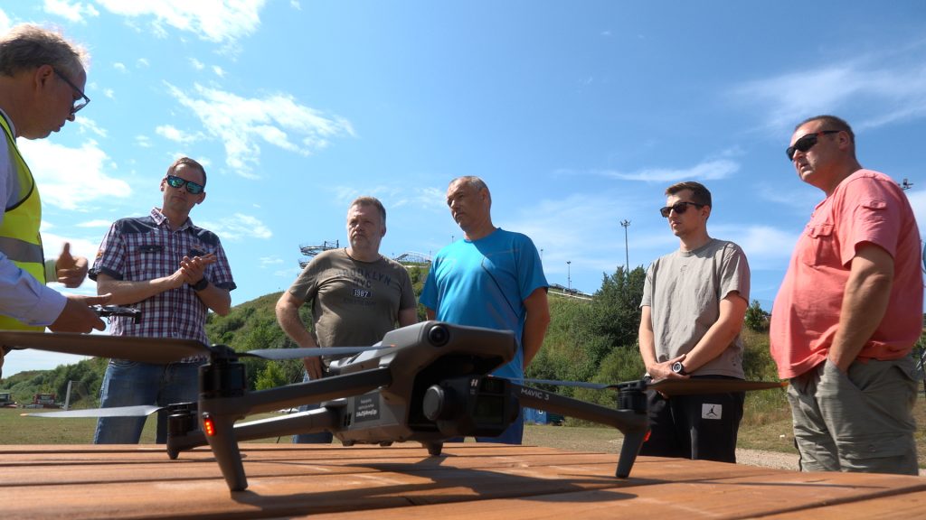 Grupa vīriešu sanākuši ap dronu "DJI Mavic 3" uz galda, klausoties instruktoru