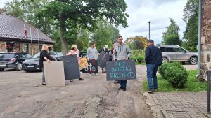 Grupa cilvēku tur plakātus ar uzrakstiem pie ielas, protestējot pret projektu