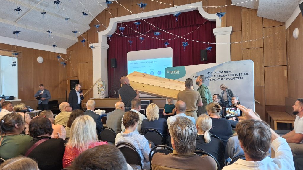 Liela cilvēku grupa sēž auditorijā, skatoties uz skatuvi, kur tiek demonstrēta koka zārka imitācija.