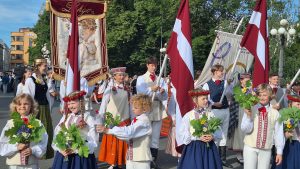 Skolēni tautastērpos ar karogiem un ziediem Dziesmu un deju svētku gājienā Rīgā.