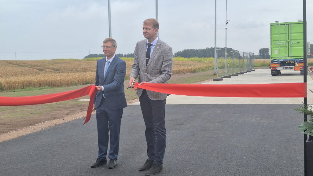 Divi vīrieši, viens uzvalkā un otrs žaketē, griež sarkanu atklāšanas lenti, atrodoties jaunā biometāna ievades punkta atklāšanā.
