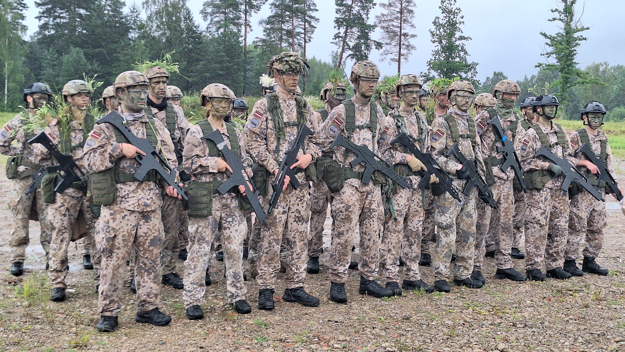 Jaunsargi militārajās uniformās ar ieročiem, stāvot ierindā ārpus telpām