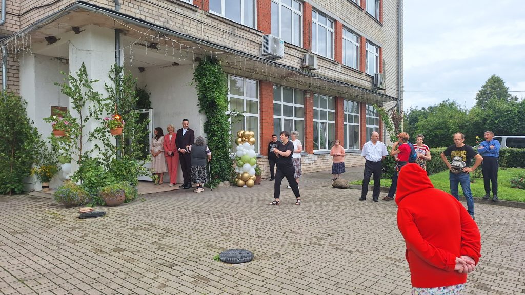 Cilvēku grupa pulcējas pie ēkas ieejas, lai atzīmētu svinīgu pasākumu