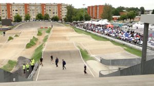 Plašs skats uz BMX sacensību trasi Valmierā ar skatītāju tribīnēm un apkārtējām ēkām.