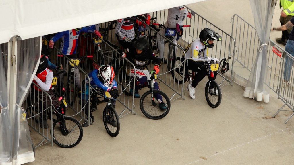 BMX braucēji stāv pie starta vārtiem, gatavojoties sacensībām zem telts.