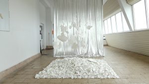 Balta porcelāna instalācija uz grīdas ar peldošiem objektiem sudrabainas drapērijas priekšā