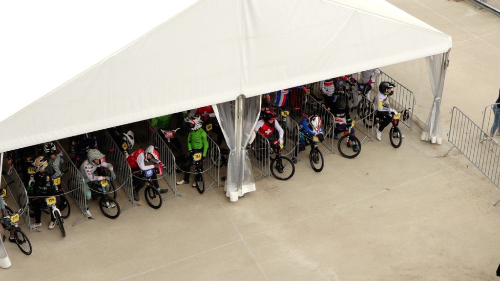 Plašs skats uz BMX braucējiem, kas gaida zem baltas telts pirms sacensību starta.