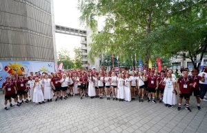 Liela jauniešu grupa, kas ir Latvijas izlase Eiropas Jauniešu Olimpiskajā festivālā, stāv brīvā dabā un pozē fotogrāfijai, turot Latvijas karodziņus.