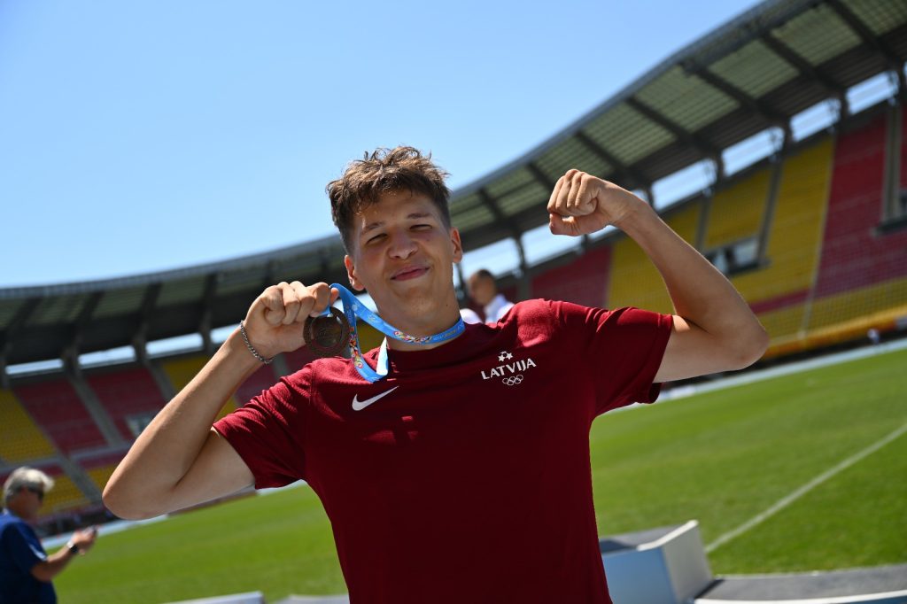 Jauns vīrietis sarkanā sporta kreklā ar medaļu ap kaklu, stāv sporta stadionā un rāda muskuļus.