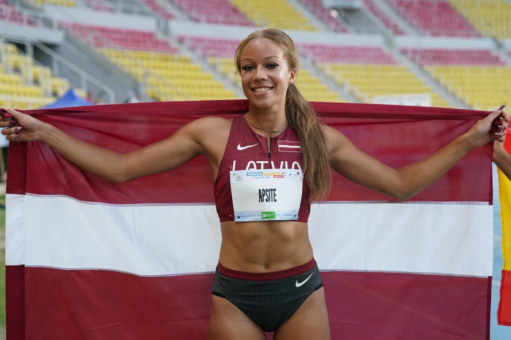 Jauna sportiste sarkanā sporta tērpā ar numuru un Latvijas karogu, smaidot stāv sporta stadionā.