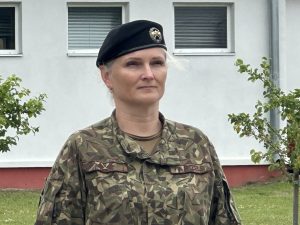 Sieviete militārā formā un beretē stāv ārā pie ēkas.