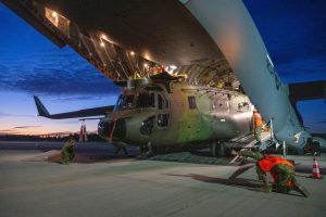Liels transporta helikopters "CH-147 Chinook" tiek izkrauts no kravas lidmašīnas tumsā, kamēr militārpersonas veic izkraušanas darbus